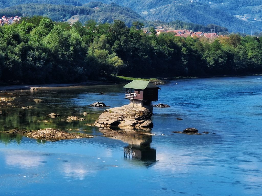 Panorama kucice na steni usred Drine sa naseljem i brdima u pozadini kod Bajine Baste