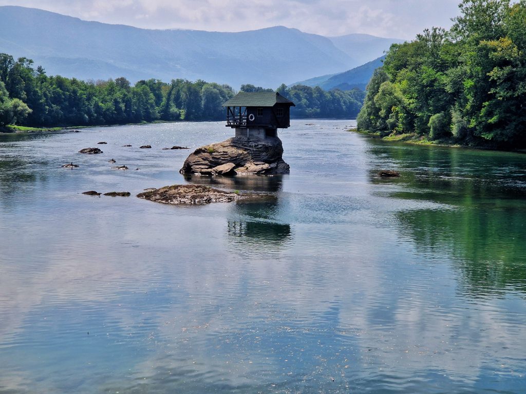 Kucica na steni usred Drine sa maglovitim planinama Tare u pozadini pod plavim nebom