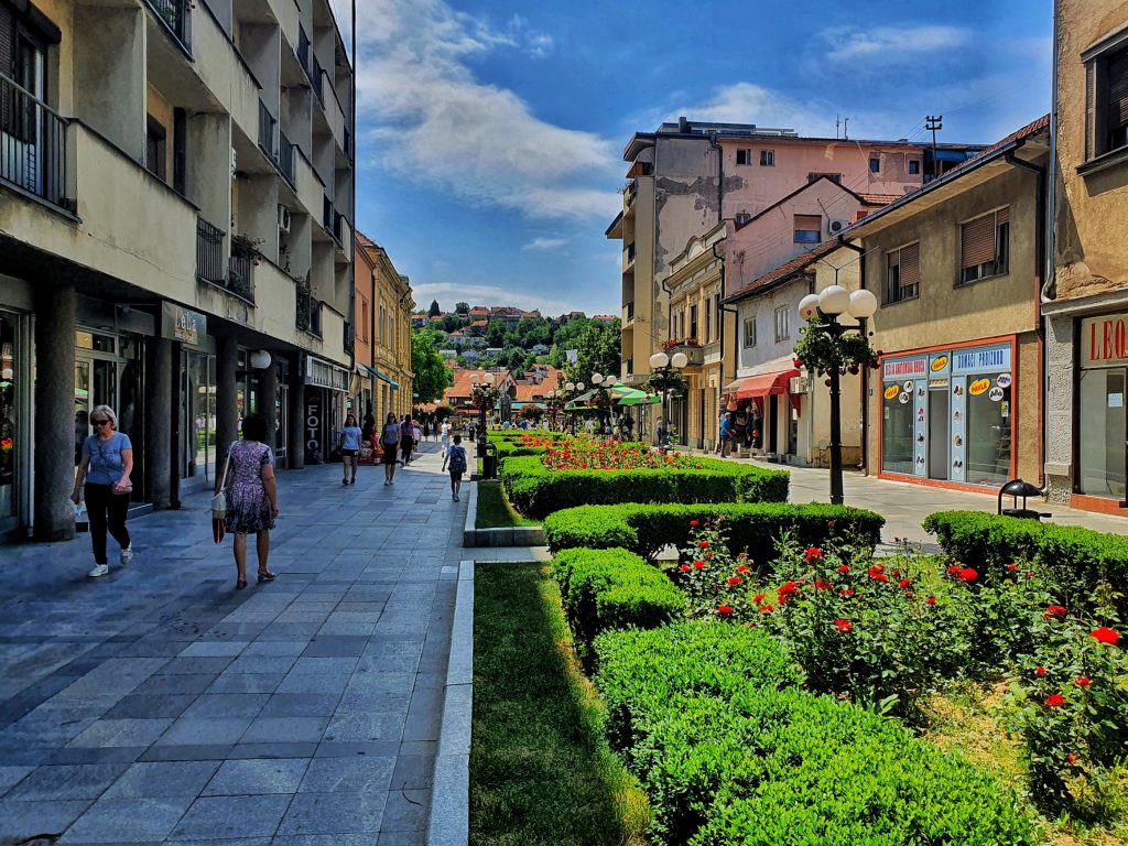 Pešačka zona u centru Valjeva, Ulica kneza Miloša sa cvetnim žardinjerama i prodavnicama pod vedrim nebom.
