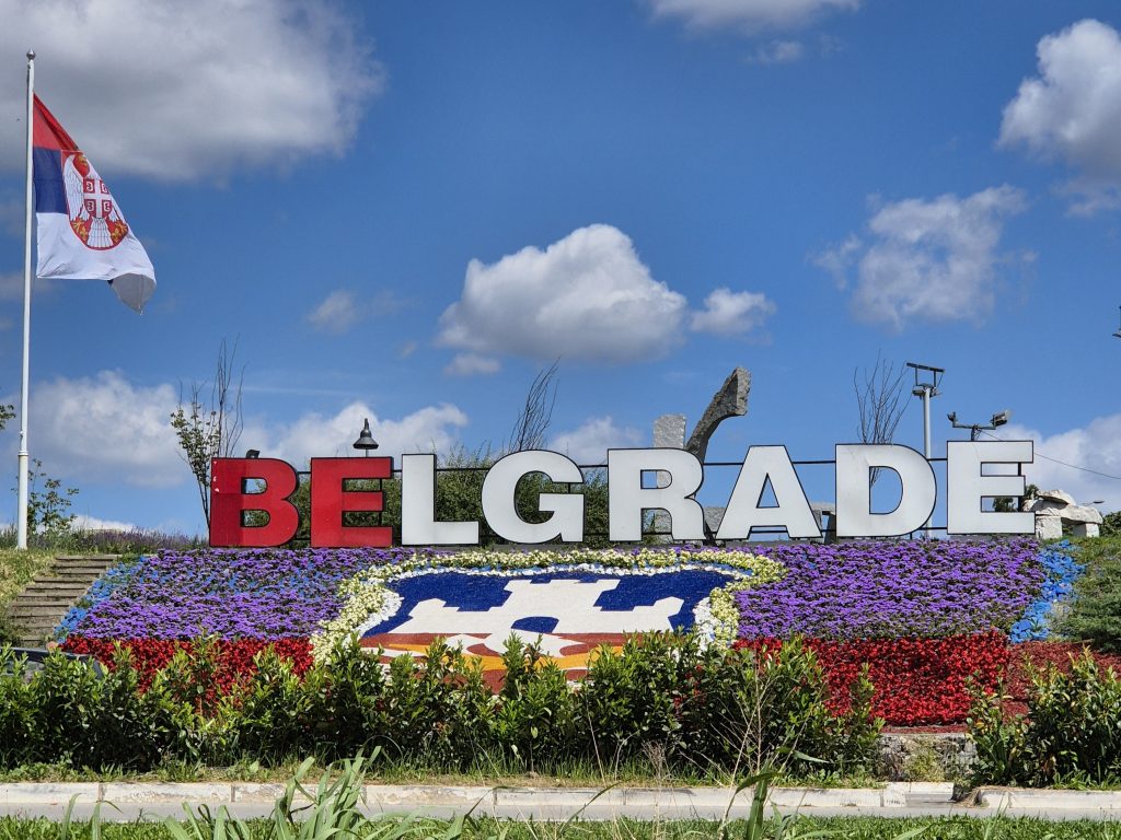 Veliki dekorativni natpis Belgrade sa grbom grada od cveca i zastavom Srbije koja se vijori na plavom nebu sa belim oblacima