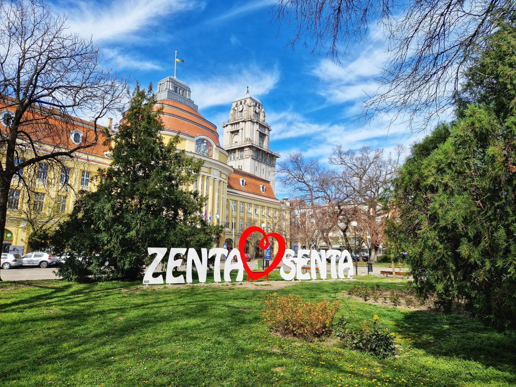 Pogled na monumentalnu žutu Gradsku kuću u Senti sa visokim tornjem, ispred koje se nalazi beli natpis Zenta-Senta sa crvenim srcem na zelenom travnjaku