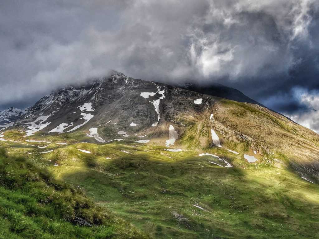 Moćni alpski vrh duz grosglokner hochalpenstrasse pod gustim tamnim olujnim oblacima i osuncanim zelenim padinama sa ostacima snega