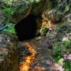 Tamni, kružni otvor starog rudarskog okna usečen u stenovitu i zemljanu padinu prekrivenu mahovinom i lišćem.