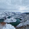 Pogled sa visine na zaleđene, smaragdno-zelene meandre reke Uvac koji se vijugaju kroz snegom prekrivene litice kanjona pod sivim zimskim nebom.