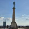 Spomenik Pobednik na visokom postolju na Kalemegdanu u Beogradu sa posetiocima na platou i modernim zgradama u daljini