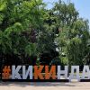 Veliki turistički natpis #KIKINDA u beloj i narandžastoj boji na popločanom trgu ispred bujnog zelenog drveća