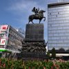 Monumentalni crni spomenik Oslobodiocima Nisa na trgu ispred visoke staklene zgrade hotela Ambasador pod plavim nebom