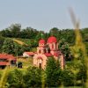 Pravoslavna crkva sa jarkocrvenim kupolama u ruralnom predelu okruzena zelenim drvecem i vocnjacima pod vedrim nebom